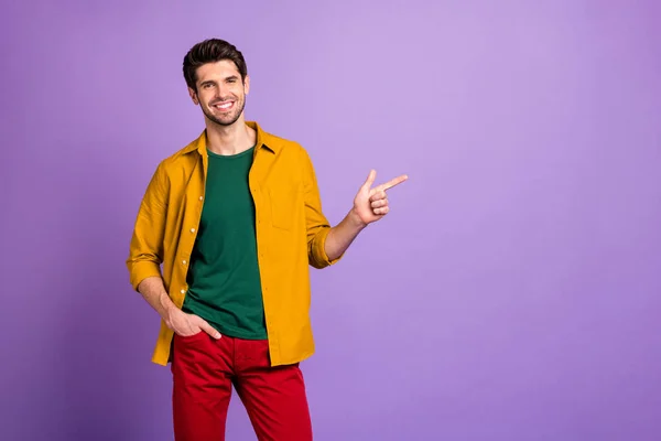Фото веселого позитивного зубастого сияющего парня, указывающего на пустое пространство в штанах, показывающего новую информацию изолированного фиолетового пастельного цвета фона — стоковое фото