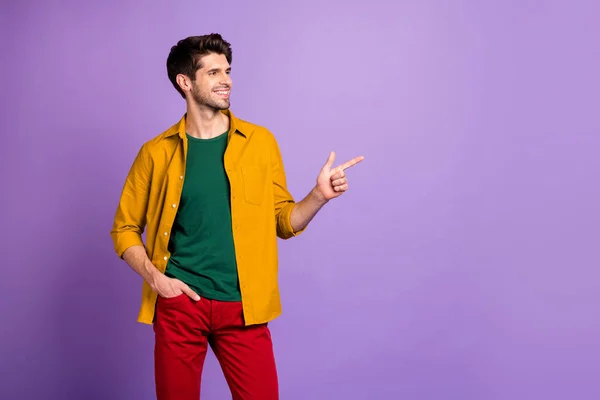Foto di allegro uomo positivo fiducioso che punta nello spazio vuoto con mano in tasca di pantaloni rossi isolato colore viola pastello sfondo — Foto Stock