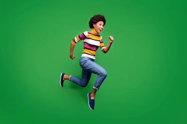 Full body profiel zijkant foto van gekke vrolijke afro-Amerikaanse meisje sprong run na de herfst kortingen voelen vreugde emoties dragen heldere kleding blauwe sneakers geïsoleerd over groene kleur achtergrond — Stockfoto