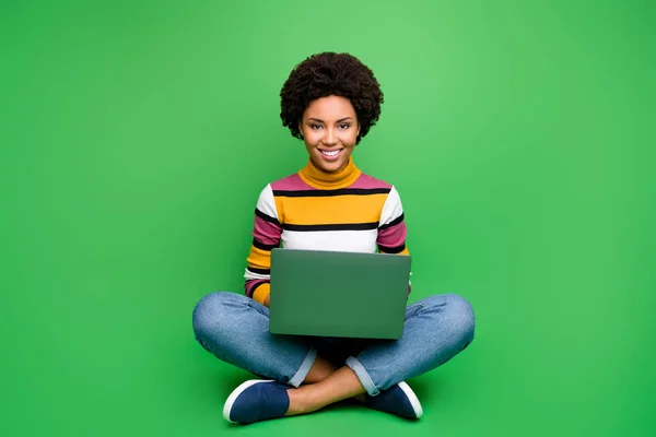Full body foto van positieve Afro-Amerikaanse meisje zitten benen gekruist gevouwen werk op computer smm werknemer concept dragen stijlvolle denim jeans outfit geïsoleerd over groene kleur achtergrond — Stockfoto