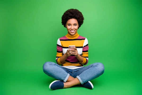 Full body photo of positive afro american girl blogger sit legs cross folded use smart phone read social network news chill chat wear shine outfit isolated green color background — Stock fotografie