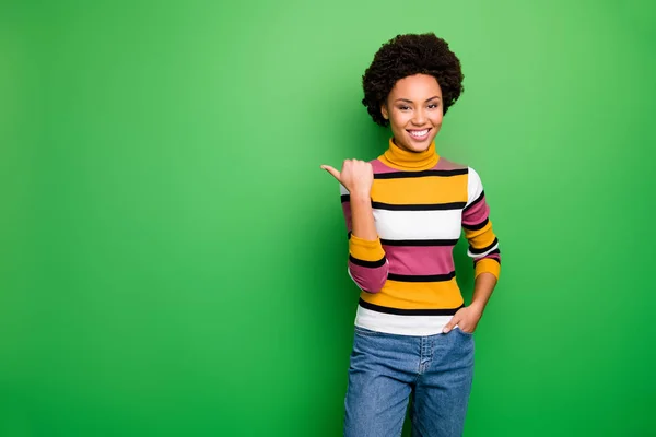 Foto di bella pelle scura ondulato signora sales manager proponendo buoni prezzi dito pollice diretto spazio vuoto usura casual pullover a righe jeans isolato colore verde sfondo — Foto Stock