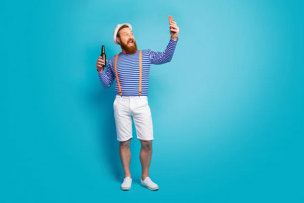 Full body foto van gekke roodharige baard man hebben vakantie ontspannen bloggen maken selfie tonen zijn fles bier dragen oranje bretels witte shorts zon hoofddeksel geïsoleerde blauwe kleur achtergrond — Stockfoto