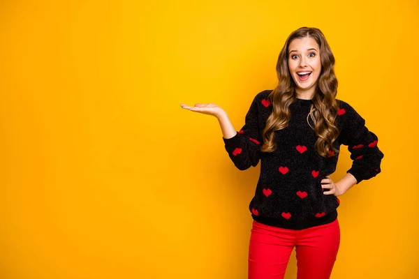 Photo of funky cheerful lady open mouth holding open palm product low season shopping prices wear hearts pattern pullover red trousers isolated yellow color background — 스톡 사진