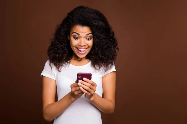 Πορτρέτο του κατάπληκτος τρελό afro american blogger κορίτσι χρησιμοποιούν κινητό τηλέφωνο πάρει κοινοποίηση κοινωνικών δικτύων ματιά κραυγή wow omg φορούν λευκό t-shirt απομονώνονται πάνω από καφέ φόντο χρώμα — Φωτογραφία Αρχείου