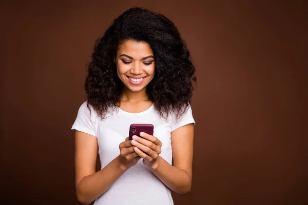 Portrét pozitivní veselý afro americká dívka použít chytrý telefon číst sociální média zprávy vzhled obrazovky nosit ležérní styl oblečení izolované přes hnědé barvy pozadí — Stock fotografie