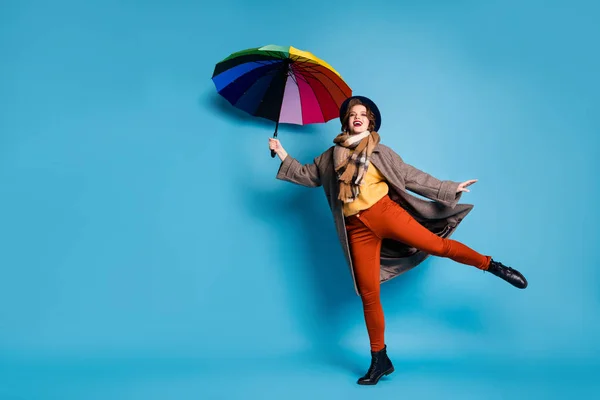 Полная длина фото смешного путешественника леди поднимая держать красочный зонтик воздуха дует износ случайные длинные серые пальто пуловер оранжевые брюки шляпа обувь изолированный синий цвет фона — стоковое фото