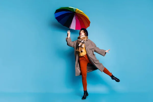 Full body foto van gekke reiziger dame opvoeden hold kleurrijke paraplu wind blazen slijtage casual lange grijze jas geel pullover oranje broek hoed schoenen geïsoleerde blauwe kleur achtergrond — Stockfoto