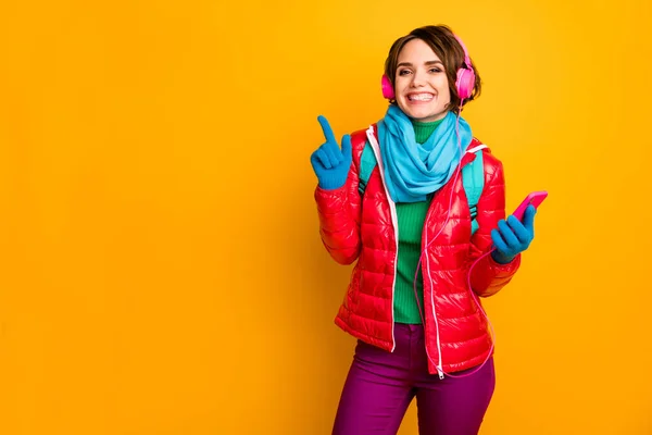 Foto von lustigen Bloggerin Dame halten Telefon hören Kopfhörer Richten Finger leer Raum tragen lässig roten Mantel blauer Schal Handschuhe Hose isoliert gelb Farbe Hintergrund — Stockfoto