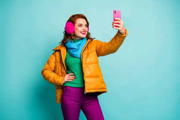 Foto von lustigen hübschen Dame halten Telefon Hände Selfies machen tragen trendige lässig Ohrenbedeckungen gelber Mantel Schal magenta Hose Pullover isoliert teal Farbe Hintergrund — Stockfoto