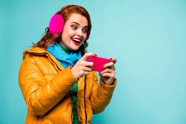 Foto av ganska rolig dam hålla telefon beroende videospel spelare öppna munnen upphetsad slitage trendig casual öronmuffar gul överrock halsduk isolerad teal färg bakgrund — Stockfoto