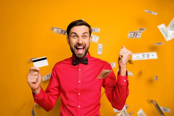 Photo of attractive funny guy hold plastic credit card rich person money bucks falling everywhere screaming wear trendy red shirt bow tie clothes isolated yellow color background — Stock Photo, Image