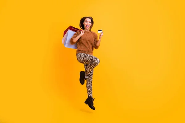 Full length profile photo of funny lady jump high carry packs shopaholic hold plastic credit card wear casual fluffy pullover leopard trousers boots isolated yellow color background — Stock Photo, Image