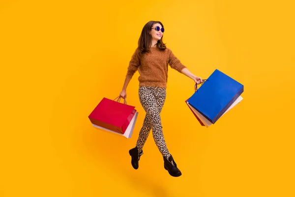 Foto de comprimento total de senhora elegante engraçado pulando alto transportar muitos pacotes de desgaste comprador animado suéter fofo leopardo calças botas isoladas cor amarela fundo — Fotografia de Stock