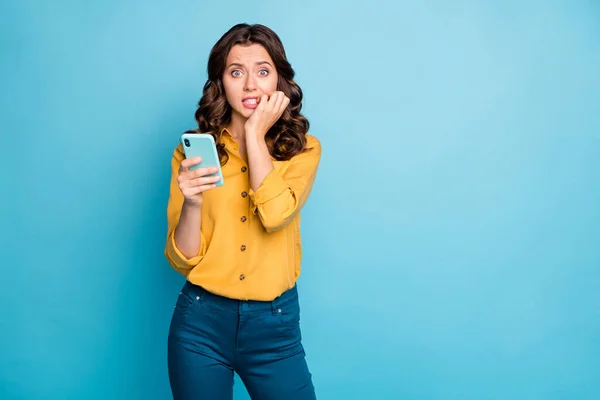 Photo of pretty lady holding telephone hands biting fingers eyes full of fear negative post comments noś żółtą koszulę spodnie odizolowane niebieski kolor tła — Zdjęcie stockowe