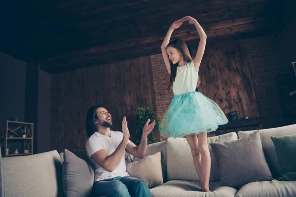 Foto av liten energisk söt dam upphetsad stilig pappa tittar dotter skolan dans höja händerna som ballerina sitter soffa klappa vapen ovation hus rum inomhus — Stockfoto