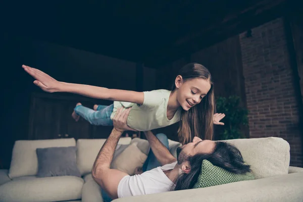Photo of little beautiful lady excited handsome daddy lying comfy sofa hold his small princess raise up air arms spread like wings pretend flight house room indoors — Stock Photo, Image