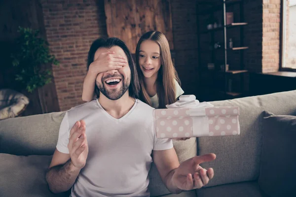 Foto av liten rolig söt dam gömmer ögonen hennes stiliga unga pappa sitta mysig soffa håller stor överraskning presentförpackning födelsedagspresent hemtrevligt rum inomhus — Stockfoto