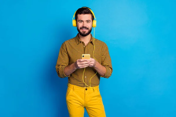 Portrait of positive cheerful guy listen music radio songs he search internet on cellphone wear shine total yellow headset checkered pants isolated over blue color background — 스톡 사진