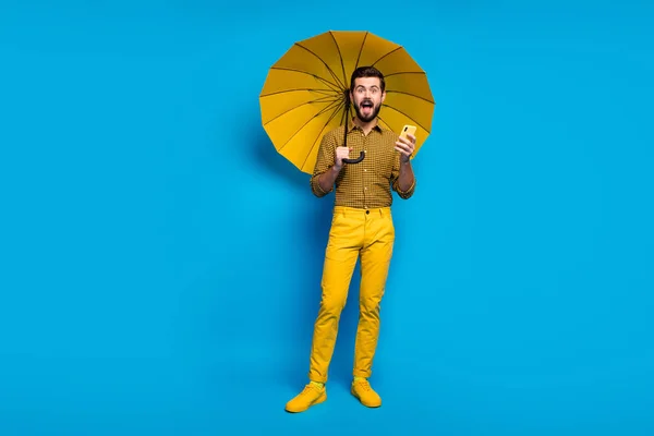 Foto de longitud completa tipo enérgico uso de teléfono inteligente obtener comentario impresionado grito omg hold proteger lluvia escudo parasol desgaste estilo elegante moda a cuadros pantalones aislados color azul fondo —  Fotos de Stock