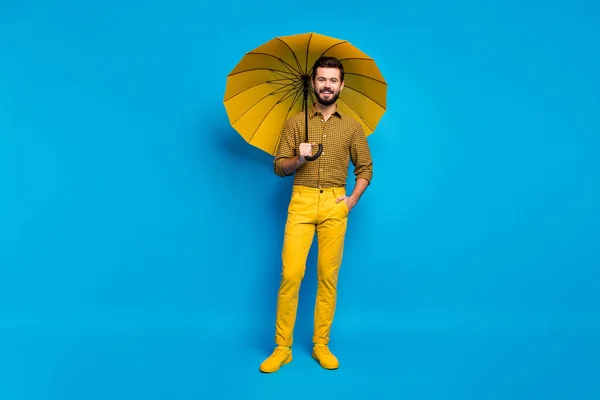 Neşeli, rüya gibi, enerjik bir adamın tam vücut fotoğrafı yağmurlu havada boş zaman, renkli parlak güneş şemsiyesi, tamamen sarı giyin mavi arka planda izole edilmiş spor ayakkabıları. — Stok fotoğraf