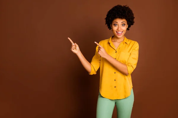 Photo of beautiful pretty excited dark skin wavy lady indicating fingers empty space sale shopping prices wear yellow shirt green trousers isolated brown color background — Stock Photo, Image