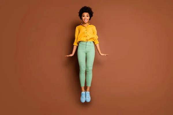 Full body photo of funny dark skin lady jump high up flirty mood toothy smiling wear yellow shirt green pants footwear isolated brown color background — Stock Photo, Image