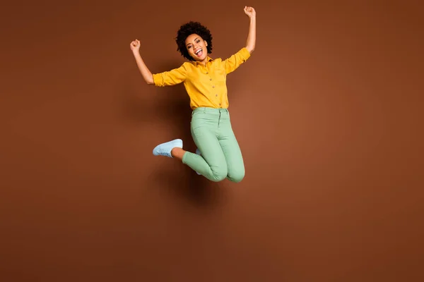 ¿Sí? Foto de cuerpo completo de la señora de piel oscura asombrada salto alto celebrando el dinero de la lotería ganar levantar los puños usar camisa amarilla pantalones verdes calzado aislado color marrón fondo —  Fotos de Stock