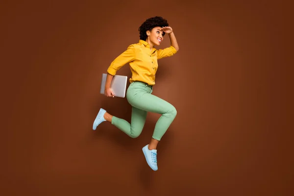 フルボディ写真のファンキーダーク肌の女性ジャンプ高ホールドノートブック急いでレッスン学校参照してください建物の近くに身に着けている黄色のシャツ緑のパンツの靴隔離された茶色の色背景 — ストック写真