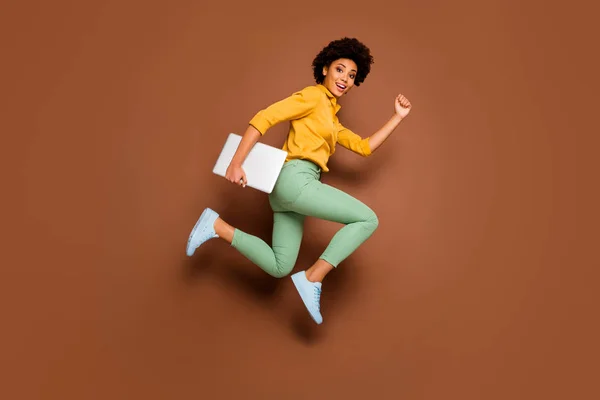 Полная длина фото фанки темная кожа леди прыгать высоко держать ноутбук спешить уроки школьница носить желтую рубашку зеленые брюки обувь изолированный коричневый цвет фона — стоковое фото