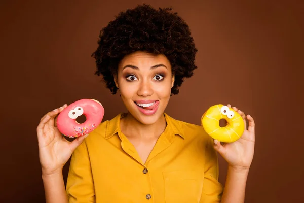 Closeup photo of funky dark skin lady hold two colorful donuts caramel eyes human faces painting hungry licking lips wear yellow shirt isolated brown color background