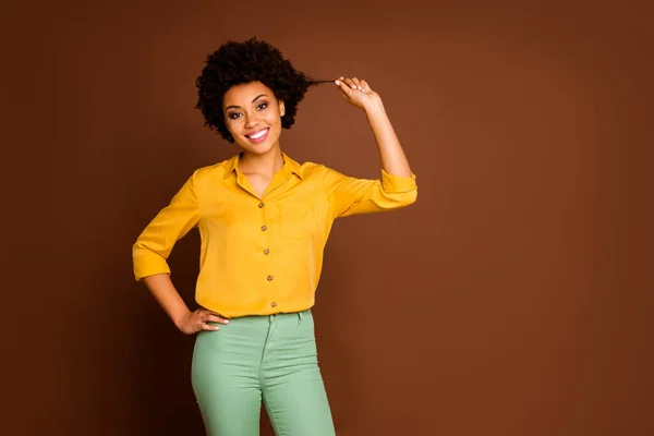 Photo of charming beautiful dark skin lady holding faliste curl pokazujące ładne wyniki po wizycie salon fryzjerski master nosić żółtą koszulę zielone spodnie odizolowane brązowy kolor tła — Zdjęcie stockowe