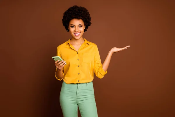 Portrait of positive cheerful afro american girl use smart phone hold hand present online adverts promotion social media sales wear yellow green outfit isolated over brown color background — 스톡 사진