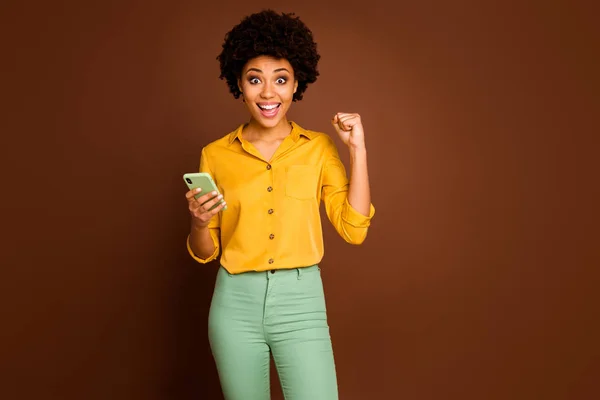 Portrait of funky lucky afro american girl student blogger use smartphone celebrate online lottery raise fists scream yeah wear yellow green pants trousers isolated brown color background