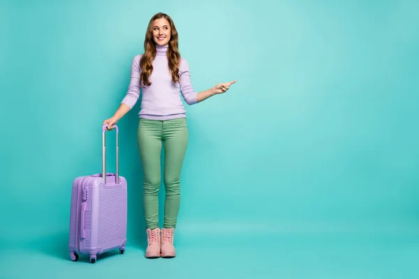 Full body foto van grappige dame luchthaven met rollende reis geval directe vinger lege ruimte kiezen beste luchtvaartmaatschappijen dragen lila trui groene broek schoeisel geïsoleerde pastel teal kleur achtergrond — Stockfoto