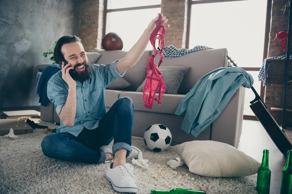 Photo of laughing hipster guy holding telephone telling friends intimacy details of his active naughty life night bad person watch red bra hands sitting floor trash after party flat indoors