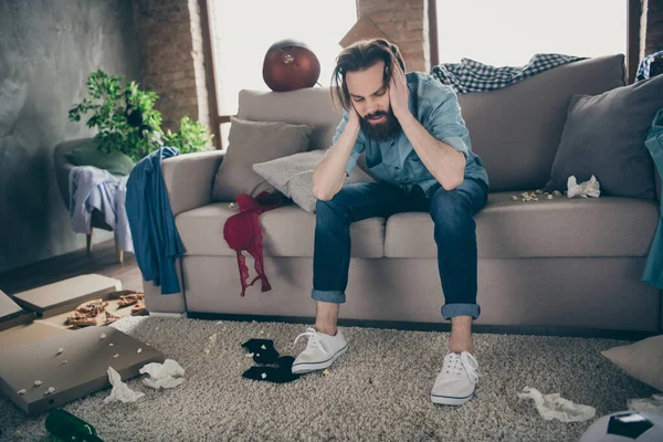 Fotografie hipster blázen chlap sedí pohovka drží hlavu opilý jídlo prádlo ležící na podlaze měl jelena strana trpí kocovina ráno bolest hlavy chaotický byt uvnitř — Stock fotografie