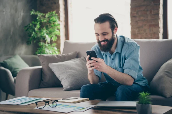 Portrait of his he nice attractive cheerful cheery brunet guy sitting on divan typing sms chatting with girlfriend at industrial loft modern style interior room house indoors — 图库照片