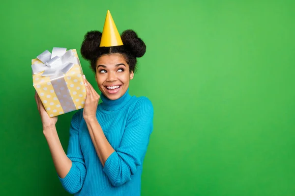 Photo of funky interested dark skin lady birthday hold big giftbox hands near ear excited to know what inside wear paper cone hat blue turtleneck isolated green color background — ストック写真