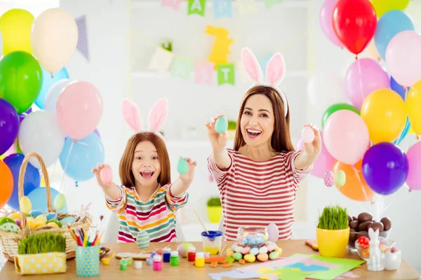 Portrait of two nice attractive glad creative cheerful cheery girls small little pre-teen daughter wearing bunny ears showing decorative eggs on sticks in white light interior room house — Stock Photo, Image