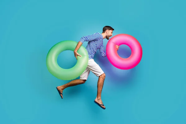 Full body profile photo of funky active guy tourist jump high rush swimming hold two colorful lifebuoys circles wear striped sailor shirt shorts flip flops isolated blue color background — ストック写真
