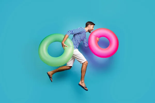 Perfil de corpo inteiro foto de cara ativa funky turista salto de alta corrida natação segurar dois colorida lifebuoys círculos desgaste listrado marinheiro camisa shorts chinelos isolado azul cor fundo — Fotografia de Stock