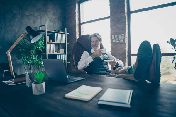 Positivo alegre aposentado homem empresa proprietário smm trabalhador sentar cadeira resto de mesa relaxar segurar caneca branca com bebida americana assistir vídeo start-up bem sucedido usar colete cinza colete no local de trabalho — Fotografia de Stock