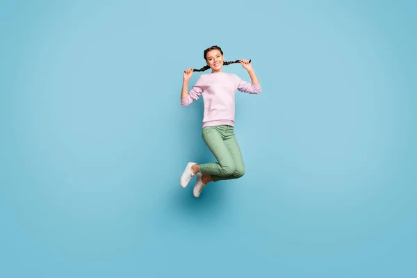 Volledige lichaam foto van grappige dame springen hoog genieten zonnige lente dag weer goed humeur houden mooie vlechten handen dragen casual roze pullover groene broek geïsoleerde blauwe kleur achtergrond — Stockfoto