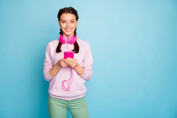 Foto de la señora increíble sosteniendo teléfono fresco auriculares brillantes escuchando radio de la juventud usuario moderno desgaste casual elegante suéter rosa pantalones verdes aislado color azul fondo — Foto de Stock