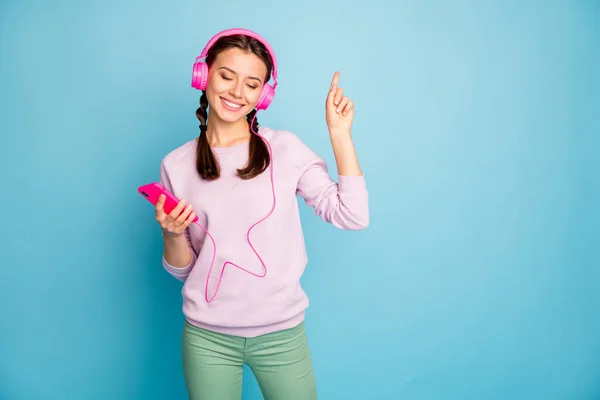 Photo of pretty lady hold telephone bright earphones on ears listen radio eyes closed raising finger rejoicing wear casual stylish pink pullover green trousers isolated blue color background