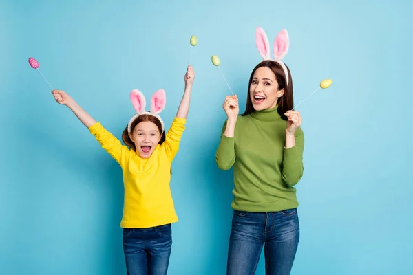 Portrait of nice attractive lovely playful cheerful cheery girls holding in hands handmade eggs on sticks having fun isolated over bright vivid shine vibrant blue color background — Stock Photo, Image