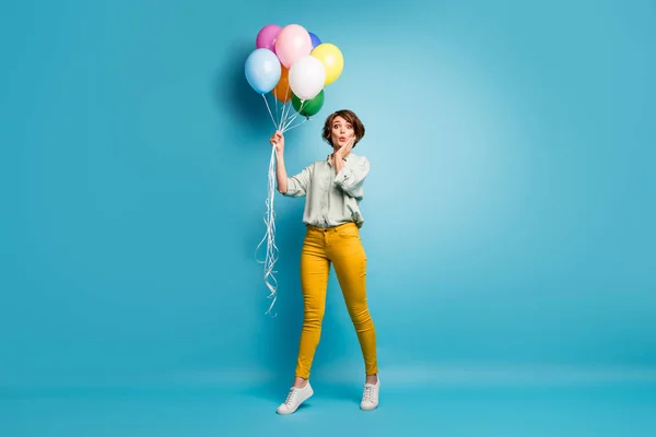 Foto von lustigen hübschen Dame zu Fuß Überraschung Geburtstagsparty halten viele Luftballons Hand auf Wange tragen lässig grünes Hemd gelbe Hose Schuhe isoliert blaue Farbe Hintergrund — Stockfoto