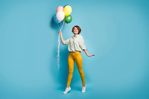 Foto de comprimento total de engraçado bonita senhora andando surpresa aniversário festa realizar muitos balões de ar usar camisa verde casual calças amarelas sapatos isolado azul cor de fundo — Fotografia de Stock