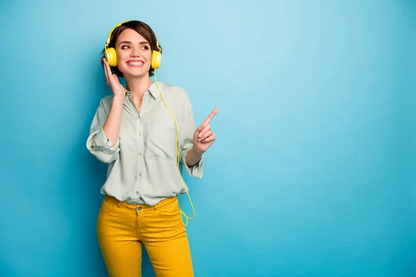 Zdjęcie wesołej pani słuchać radia stylowe słuchawki podnieść palec wskazując puste miejsce nosić zwykłą zieloną koszulę żółte spodnie odizolowane niebieski kolor tła — Zdjęcie stockowe